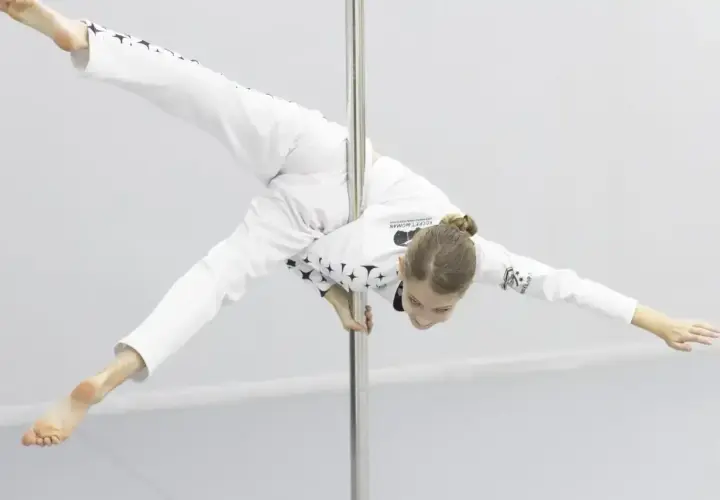 Dziewczyna w białym stroju z gwiazdami wykonuje akrobatyczną figurę na rurze. Czy pole dance to grzech? Ta młoda sportsmenka udowadnia, że to sztuka.