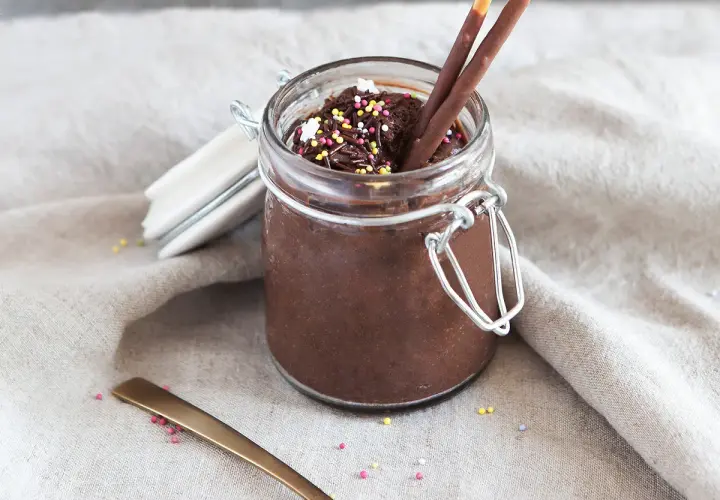 Un pot de mousse au chocolat mascarpone, garni de vermicelles colorés et de bâtonnets au chocolat. Une cuillère dorée repose à côté.