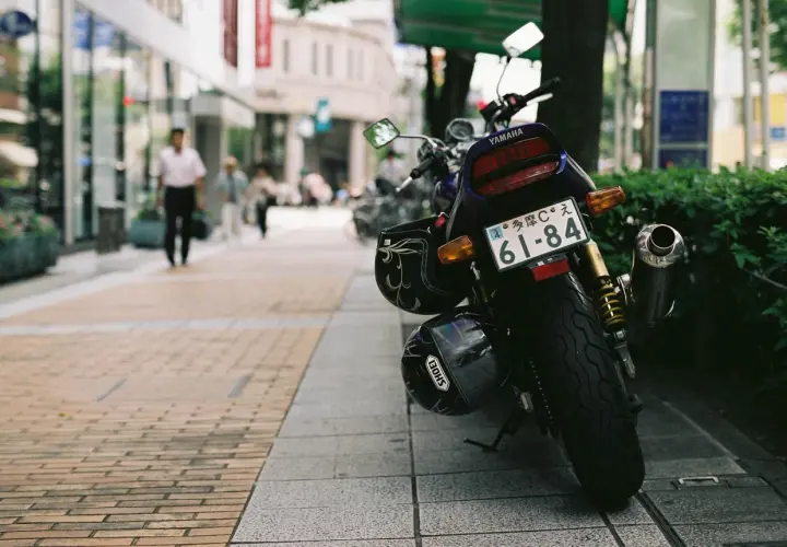 Una moto Yamaha aparcada en la acera, con dos cascos colgados. Al fondo, gente pasea por la calle.