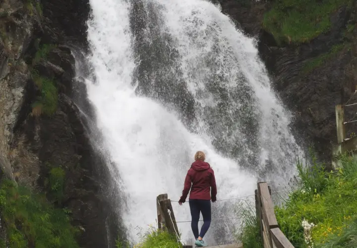 Mujer contemplando una cascada en Vielha e Mijaran. El agua cae con fuerza sobre rocas oscuras, rodeada de vegetación verde.