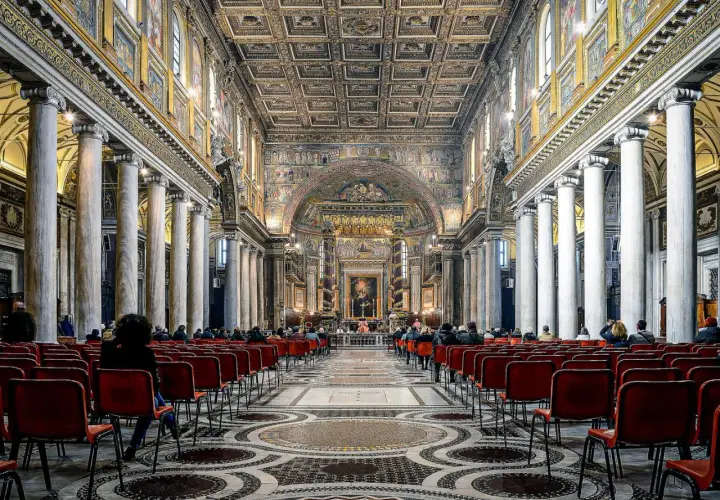Intérieur de Sainte-Marie-Majeure, Rome. Des fidèles assis sur des chaises rouges, sous des colonnes imposantes et un plafond orné.