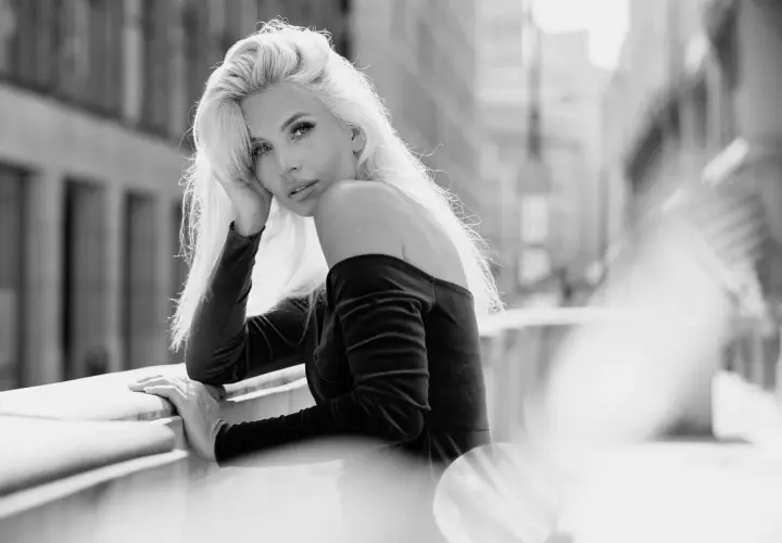 Elegancki portret w plenerze. Kobieta z blond włosami opiera się o balustradę, patrząc w obiektyw.
