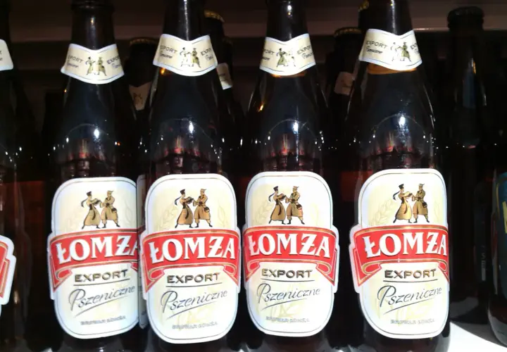 Rząd butelek piwa Łomża Export Pszeniczne. Etykiety ozdobione postaciami w strojach ludowych.