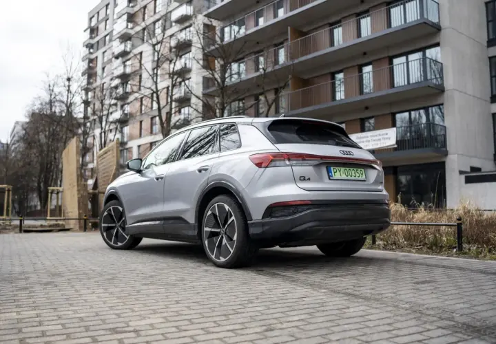 Srebrny Audi Q4 elektryk z zieloną tablicą rejestracyjną stoi na brukowanym podjeździe przed nowoczesnym budynkiem mieszkalnym.