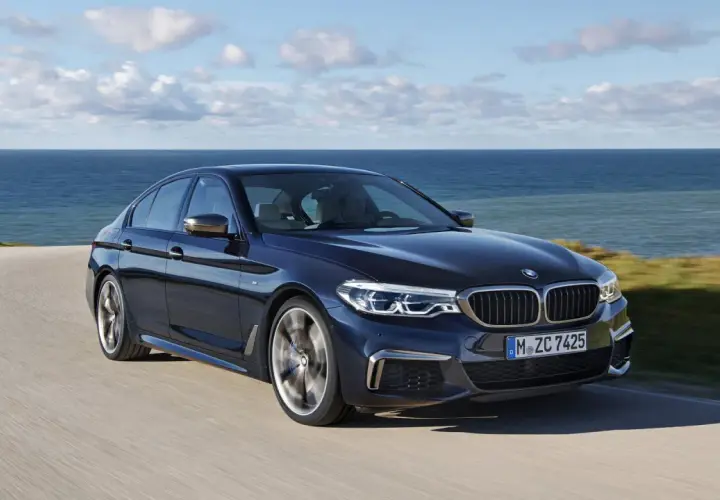 Ciemnoniebieskie BMW M550i sunie po krętej drodze nad brzegiem morza.