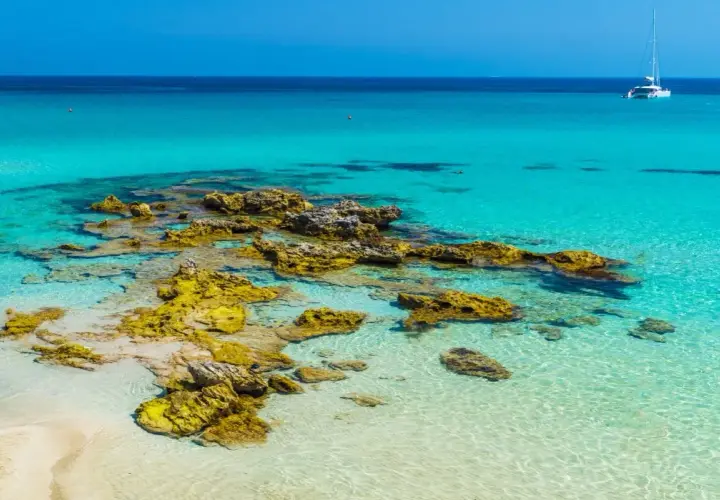 Eaux turquoise et rochers dorés sur une plage du sud de l'Italie. Un voilier blanc navigue au loin.