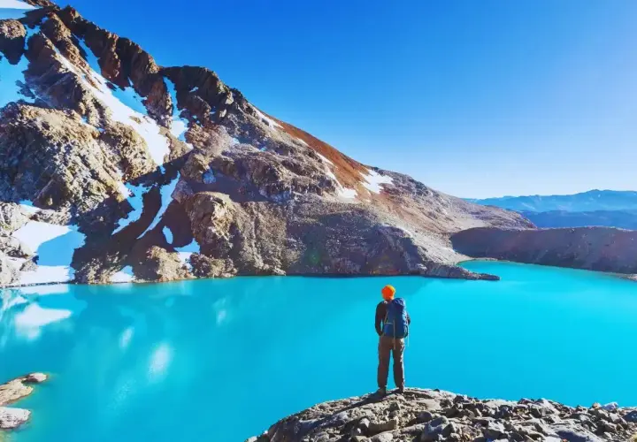 Sender solitario contempla un lago turquesa en la patagonia chilena, rodeado de montañas nevadas y un cielo azul intenso.