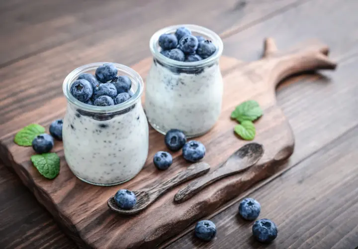 Dwa słoiczki z deserem pudding chia, ozdobione świeżymi borówkami i listkami mięty, na drewnianej desce.
