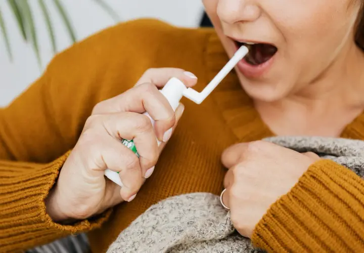 Une personne utilise un spray buccal pour soulager un mal de gorge. Elle tient le flacon blanc et dirige l'embout vers sa bouche ouverte.