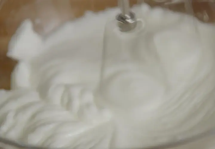 Un batteur électrique fouette un œuf blanc en neige dans un bol en verre, créant une texture aérienne et mousseuse.