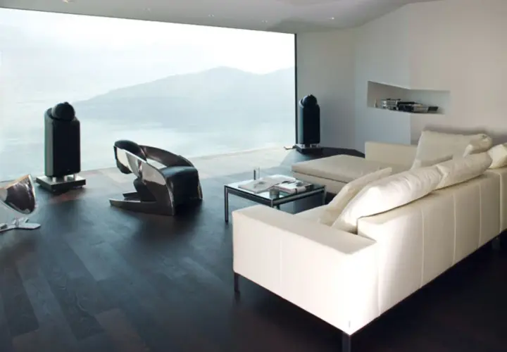 Modernes Wohnzimmer mit Panoramafenster, das eine atemberaubende Aussicht bietet. Ein weißes Ledersofa, ein futuristischer Sessel und zwei schlanke Lautsprecher ergänzen die minimalistische Einrichtung. Das Panoramafenster dekorieren wir hier mit der N...