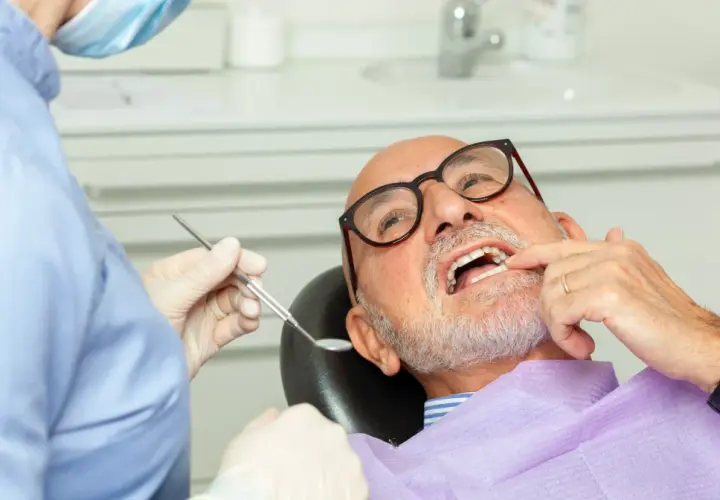 Dentysta w masce i rękawiczkach bada uśmiech starszego mężczyzny w okularach.