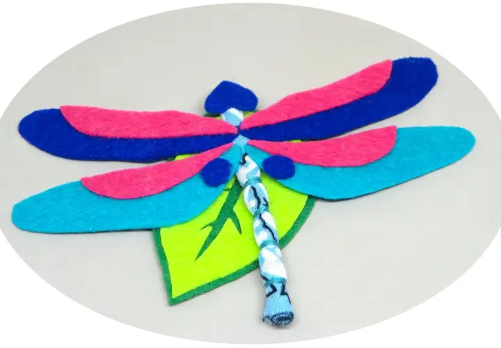 Libélula de fieltro, un proyecto de manualidades con alas azules, rosas y turquesas, posada sobre una hoja verde.