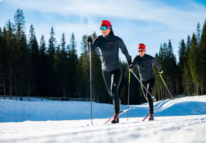 Dwie kobiety w sportowych strojach pokonują trasę narciarską. Długość nart biegowych jest kluczowa dla ich szybkości i stabilności na śniegu.