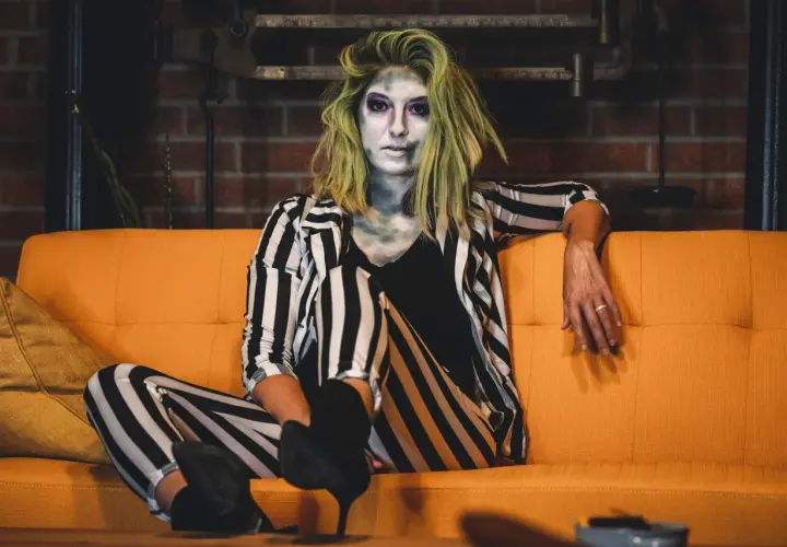 Donna con trucco spettrale e capelli verdi, indossa un vestito da strega fai da te a righe bianche e nere, seduta su un divano arancione.