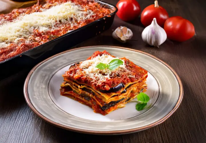 Una fetta di lasagne vegetariane, con melanzane e pomodoro, guarnita con basilico e parmigiano. Sullo sfondo, ingredienti freschi.
