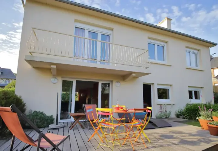 Maison divisée en 2 logements avec terrasse en bois, mobilier coloré et transat. Jardin verdoyant et ciel bleu.