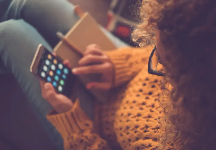 Kobieta w okularach i swetrze trzyma telefon z ikonami aplikacji i notatnik.