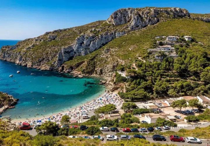 Bahía con aguas turquesas y playa abarrotada, rodeada de acantilados y vegetación. Un paraíso entre las mejores playas de Alicante.