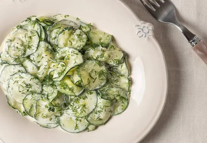 Ensalada alemana de pepino en rodajas finas, cubierta con una cremosa salsa y eneldo fresco, servida en un plato blanco.