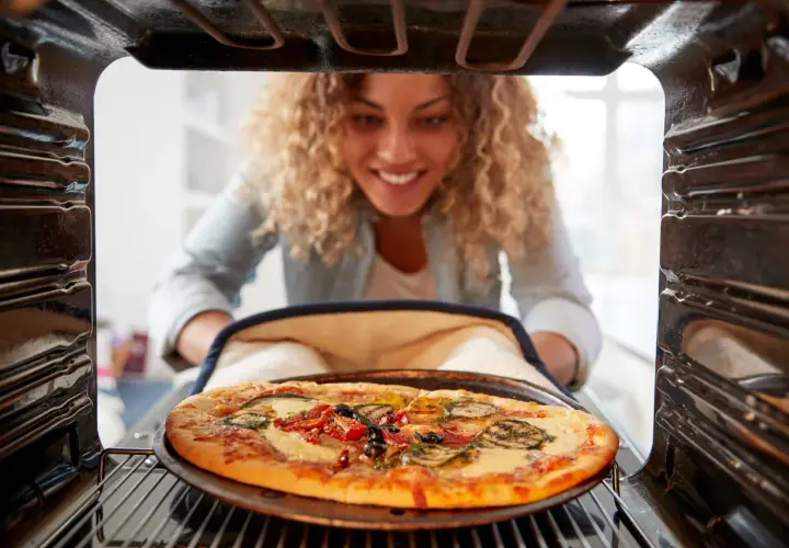 Uśmiechnięta kobieta wyjmuje z piekarnika pyszną pizzę. Widać, na jakim programie piec pizze, bo jest idealnie wypieczona.