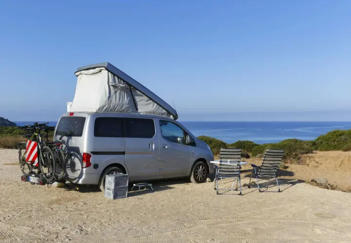 Silberner Camper mit Dachzelt und Fahrrädern am Heck, zwei Liegestühle und ein Tisch stehen bereit für entspanntes italien wildcampen am Meer.