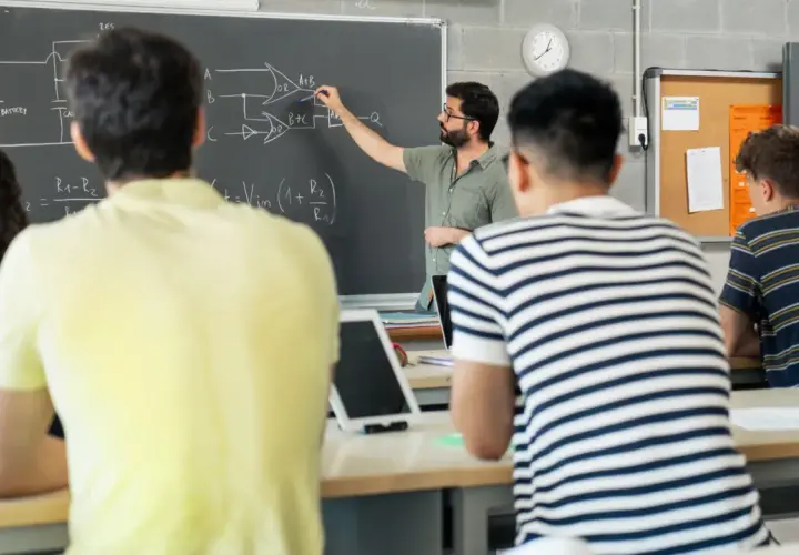 Nauczyciel tłumaczy schemat na tablicy, studenci słuchają. To część jego pensum, by przekazać wiedzę.