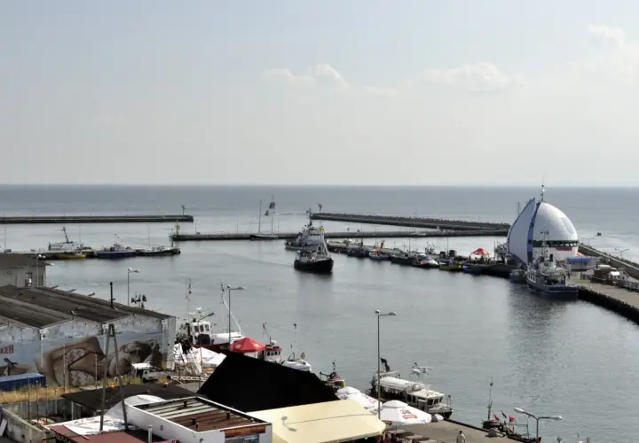 Widok na port Hel z licznymi statkami i łodziami zacumowanymi przy nabrzeżu. W tle widać morze i falochrony.