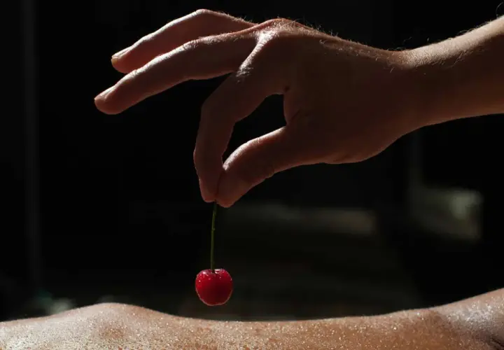 Dłoń trzyma wisienkę na cienkiej łodyżce, tuż nad wilgotną skórą. To jakby smakowanie życia na krawędzi, w znaczeniu "edging meaning".