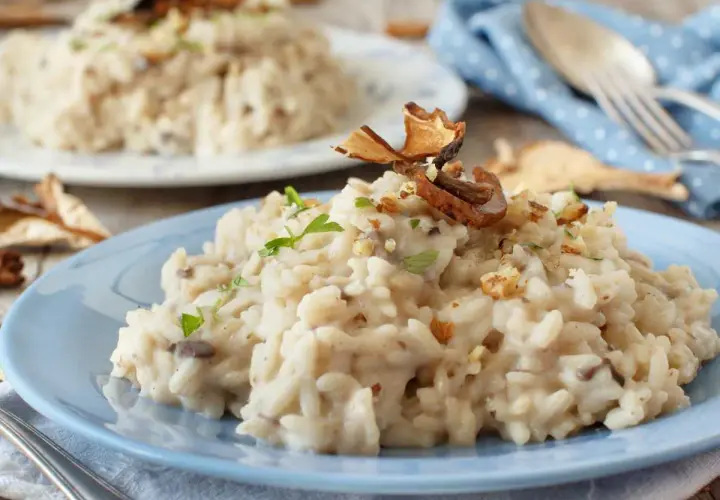 Delicioso risotto de setas cremoso, adornado con trozos de champiñones secos y nueces picadas, servido en un plato azul.