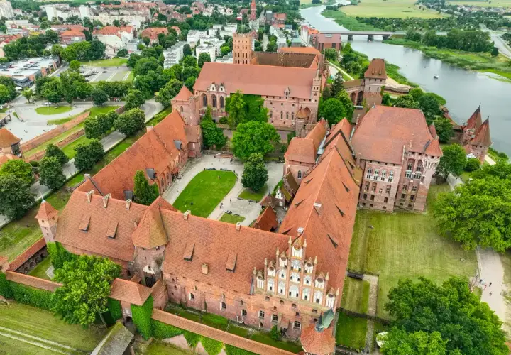 Potężny zamek w Malborku, z czerwonymi dachami i zielonymi dziedzińcami, rozciąga się wzdłuż rzeki Nogat.