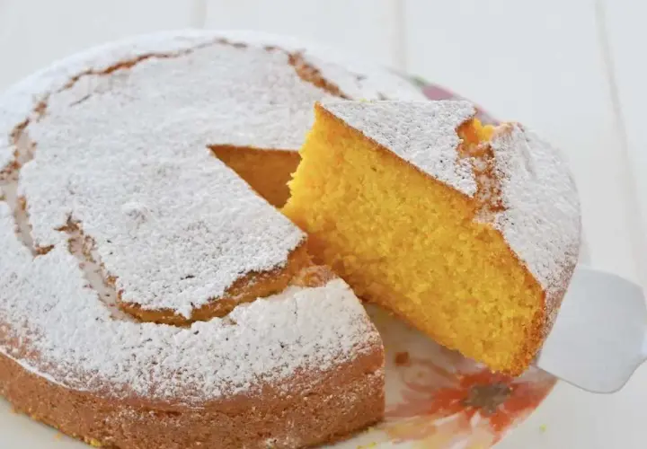 Una fetta di torta di carote e yogurt, soffice e dorata, viene sollevata da una spatola. La torta è cosparsa di zucchero a velo.