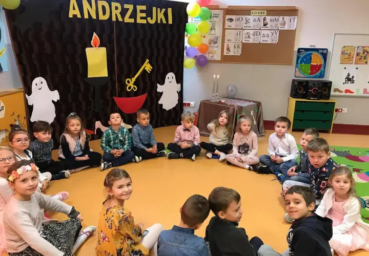 Dzieci na andrzejkach w przedszkolu, siedzą w kółku. W tle dekoracje z napisem "ANDRZEJKI", świece i duchy.