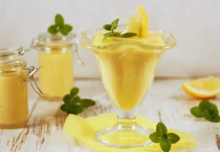 Pyszny, kremowy lemon curd w pucharku, ozdobiony plasterkiem cytryny i miętą. Obok dwa słoiczki z tym samym musem.