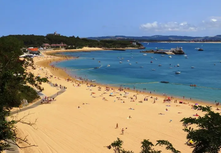 La **playa de los peligros** se llena de gente disfrutando del sol y el mar azul, con barcos anclados y edificios en la costa.