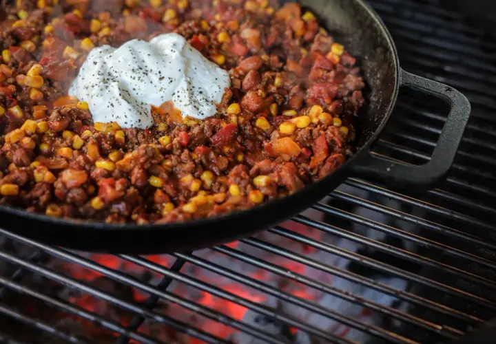 Chili con carne fumant dans une poêle sur un barbecue, garni de crème fraîche.