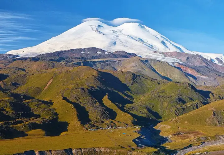 Elbrus szczyt w chmurach, otoczony zielonymi zboczami i obozem u podnóża.