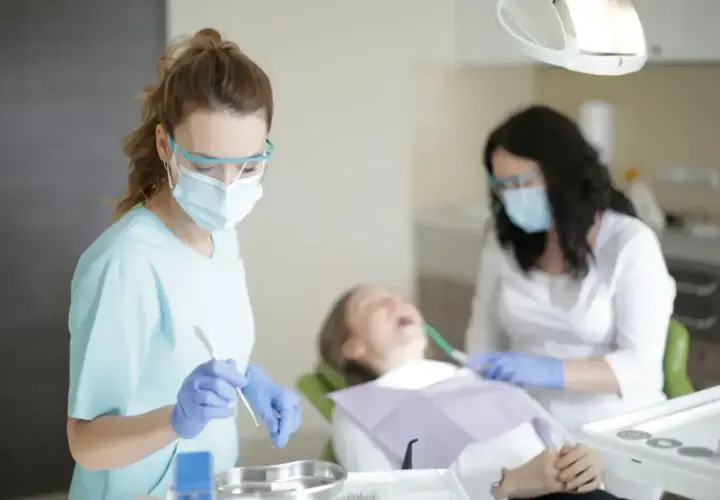 Dwie dentystki w maskach i okularach pracują przy pacjencie. Jedna z nich trzyma narzędzie, być może do usuwania pozostawionych szwów nierozpuszczalnych.