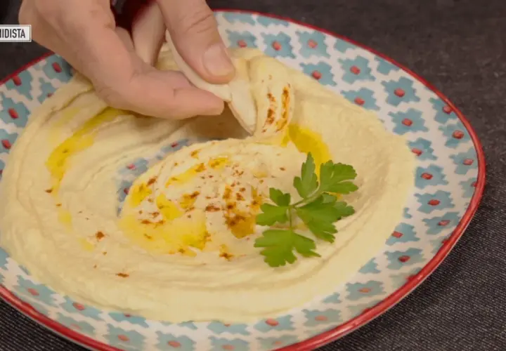 Un trozo de pan pita se sumerge en un cremoso hummus, adornado con aceite de oliva y perejil. Descubre qué es el tahini y cómo prepararlo.