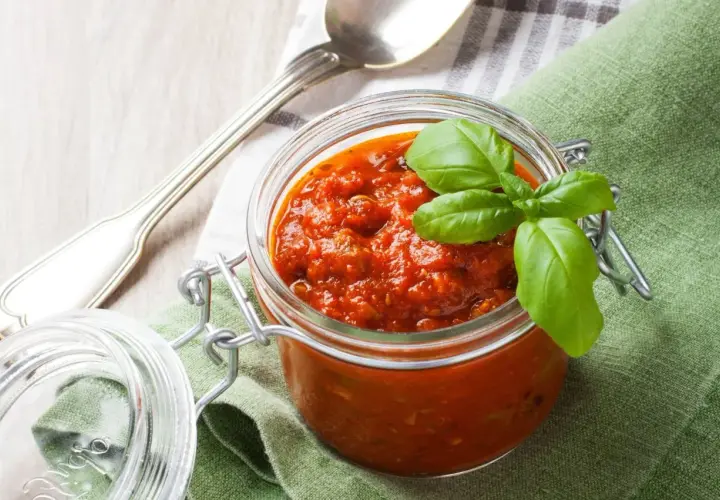 Tarro de cristal con salsa napolitana casera, decorado con hojas de albahaca fresca. Cuchara plateada y servilleta verde al lado.