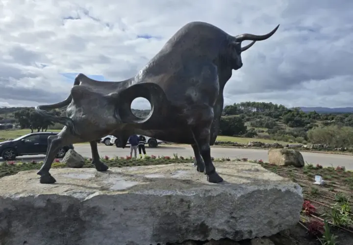 Escultura de toro en la plaza toros Valdemorillo. Paisaje rural con colinas verdes y cielo nublado.