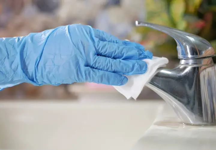 A gloved hand wipes a chrome faucet with a white cloth.