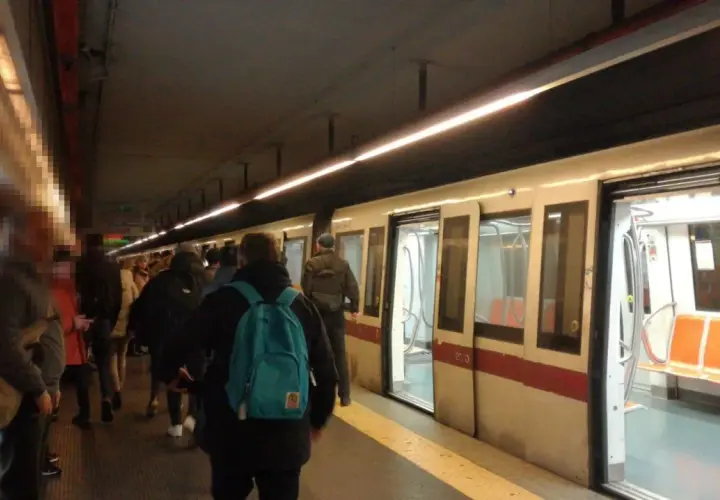 Persone in attesa sul binario della metro domenica, con un treno bianco e rosso fermo con le porte aperte.