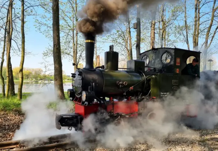Parowóz z kotłem i kominem wydzielający dym, jeden z zabytkowych lokomotyw w Polsce, na tle jeziora i drzew.