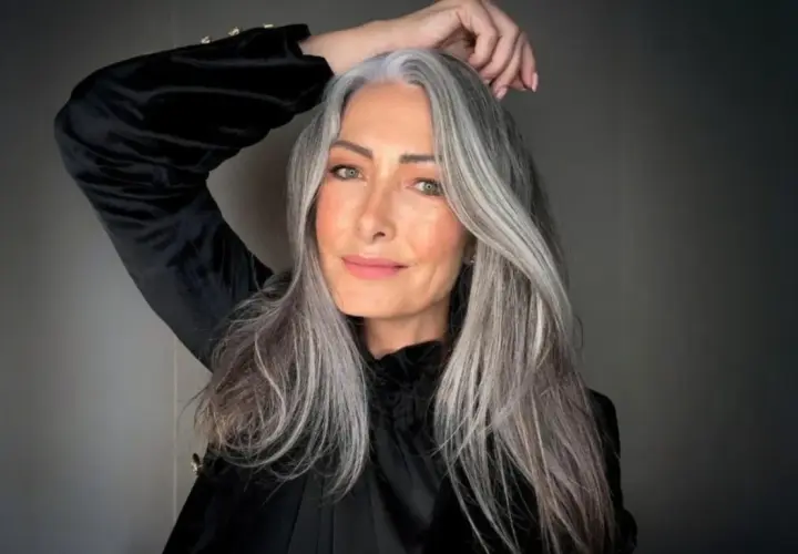 Mujer con cabello largo y ondulado, mostrando una hermosa integración de canas. Su mano acaricia su cabeza, resaltando la textura y el color natural.