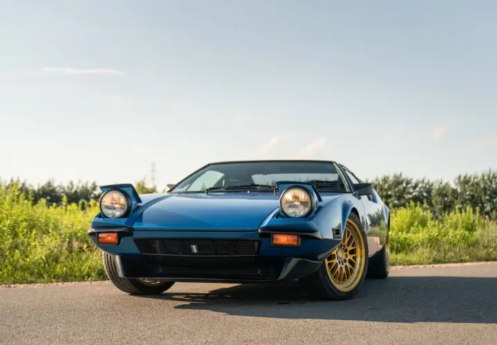 Niebieski De Tomaso Pantera z wysuwanymi światłami i złotymi felgami na tle zieleni i błękitnego nieba.