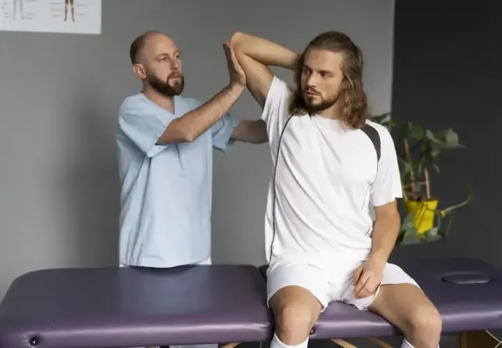 Fizjoterapeuta bada naciągnięty bark młodego sportowca siedzącego na kozetce.