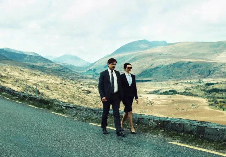 Coppia in abito cammina su strada panoramica, paesaggio montuoso. Un'immagine che evoca la recensione di "The Lobster".