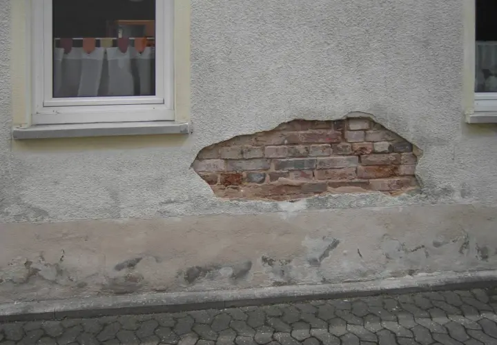 Putz löst sich im Sockelbereich, darunter Ziegelmauerwerk. Anzeichen für aufsteigende Feuchtigkeit im Sockelbereich.