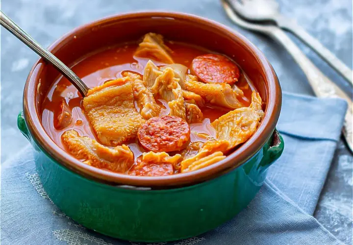Un tazón de barro con callos a la madrileña, trozos de chorizo y un caldo rojizo. Una cuchara metálica está dentro.